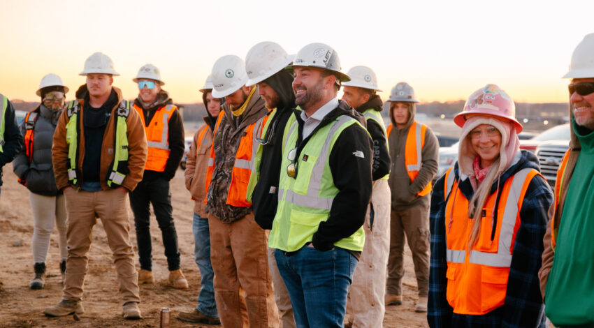Construction workers in safety gear stand on a dirt lot at dawn.
