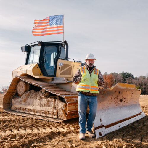 Construction worker in safety gear stands before bulldozer with flag.