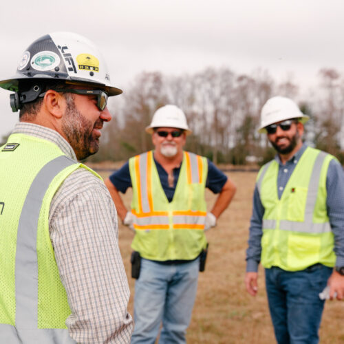 Three construction workers in safety gear smile and chat outside.