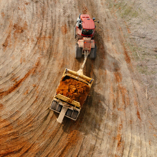 Red tractor pulls earth scraper, leaving tracks on dirt site.